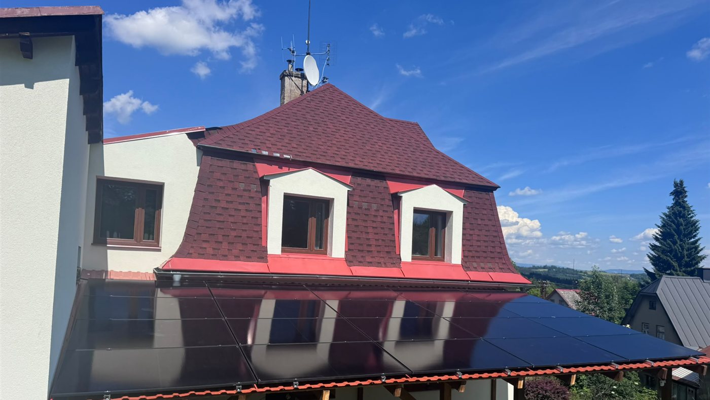 Dům s mansardovou střechou a černými panely na střeše terasy, pohled za slunečného dne